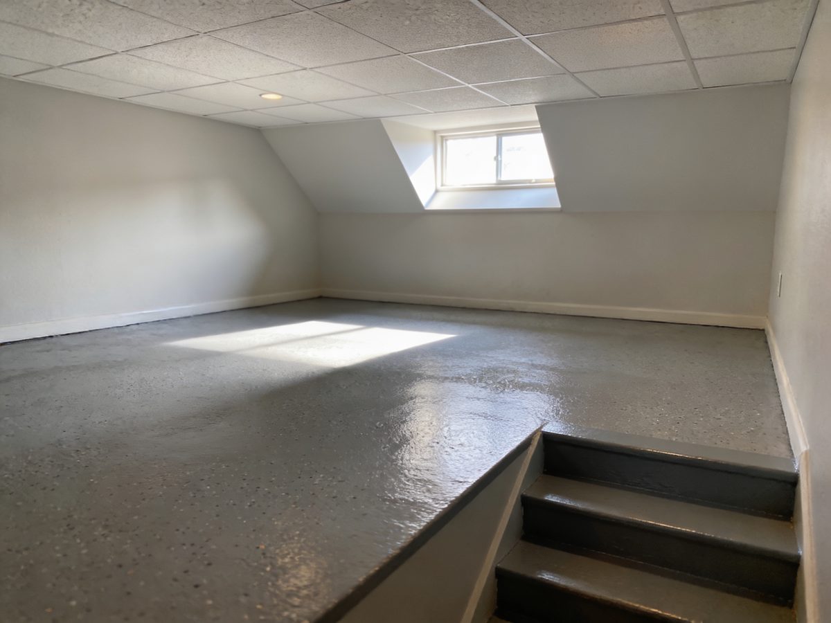 A finished basement family room with a warm grey flake floor coating, a painted stair and a window well throwing daylight across the floor